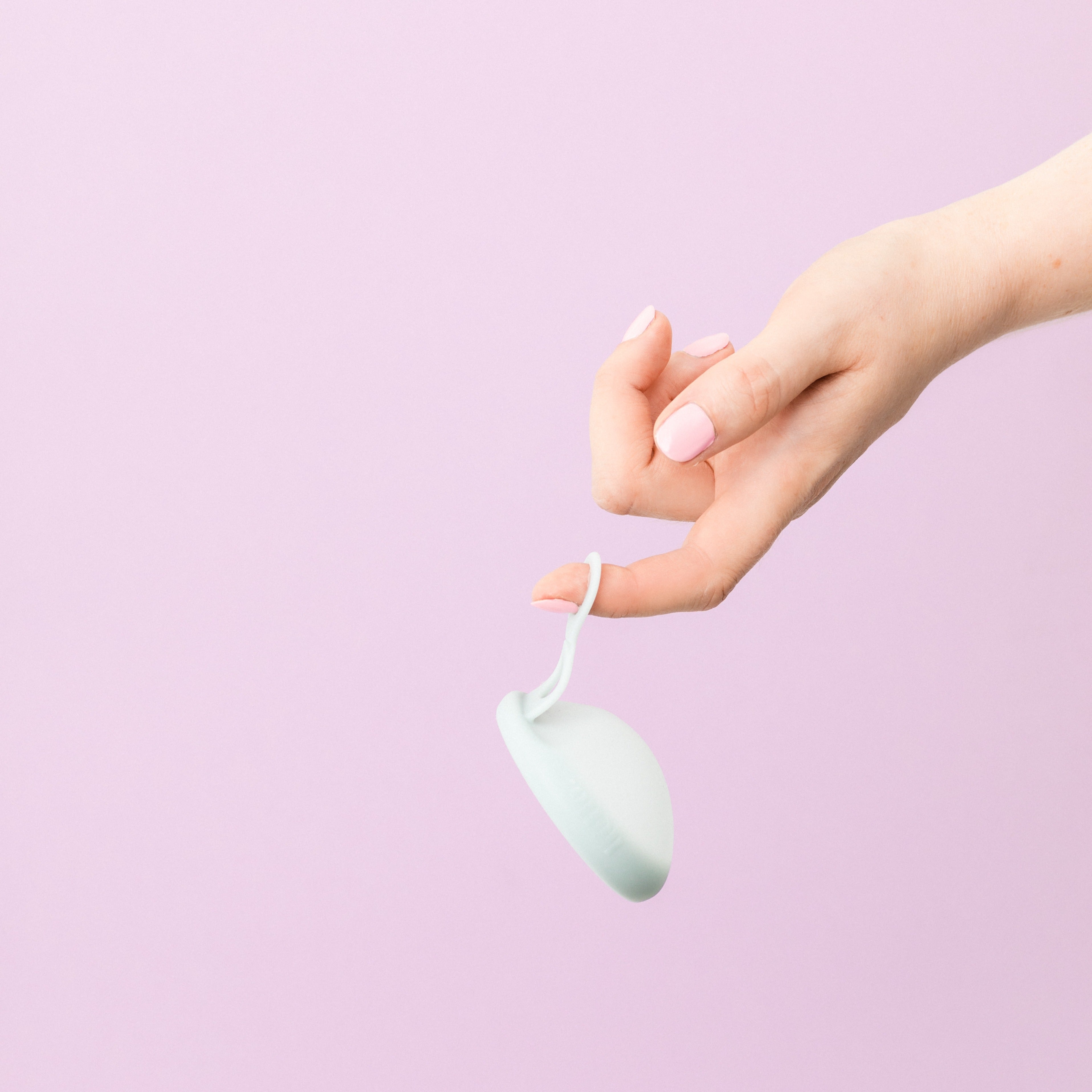 Hello Disc menstrual disc with patented double loop tabs for easier removal.Hand with pink fingernails with a finger through the bottom loop of Hello Period's menstrual disc in Sea Fog colour on a lilac background