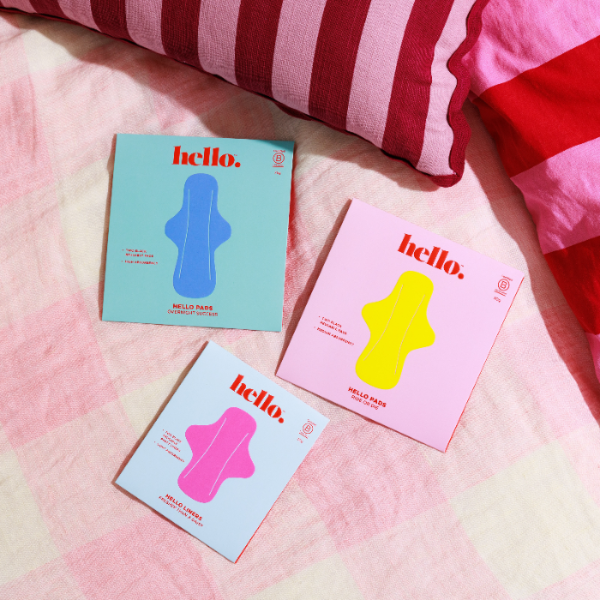 Three colourful boxes of Hello Period pads on a pink gingham bedspread with red striped pillows in the background