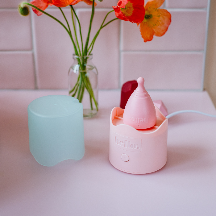 a pink bathroom with tiled wall and pink cup steriliser with a pink menstrual cup inside. a vase of orange flowers in the background. 