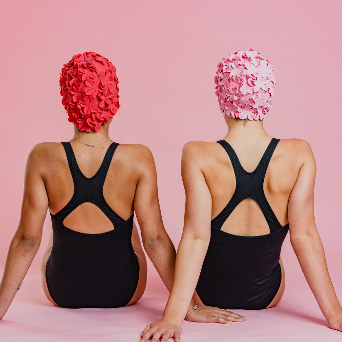 Two girls in swim caps sitting on the ground in black cross back period swimsuits facing away from the camera.