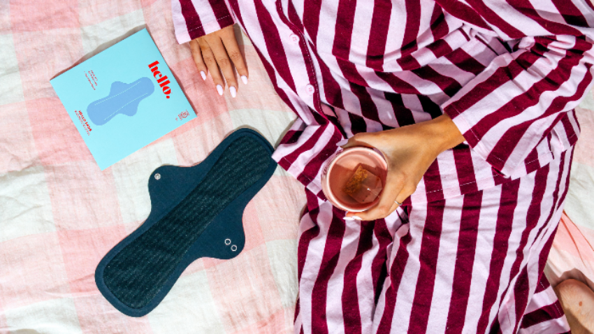 Person in a striped pyjamas robe holding a mug on a bed with a blue pad packaging, and a black Overnight Success sanitary pad.