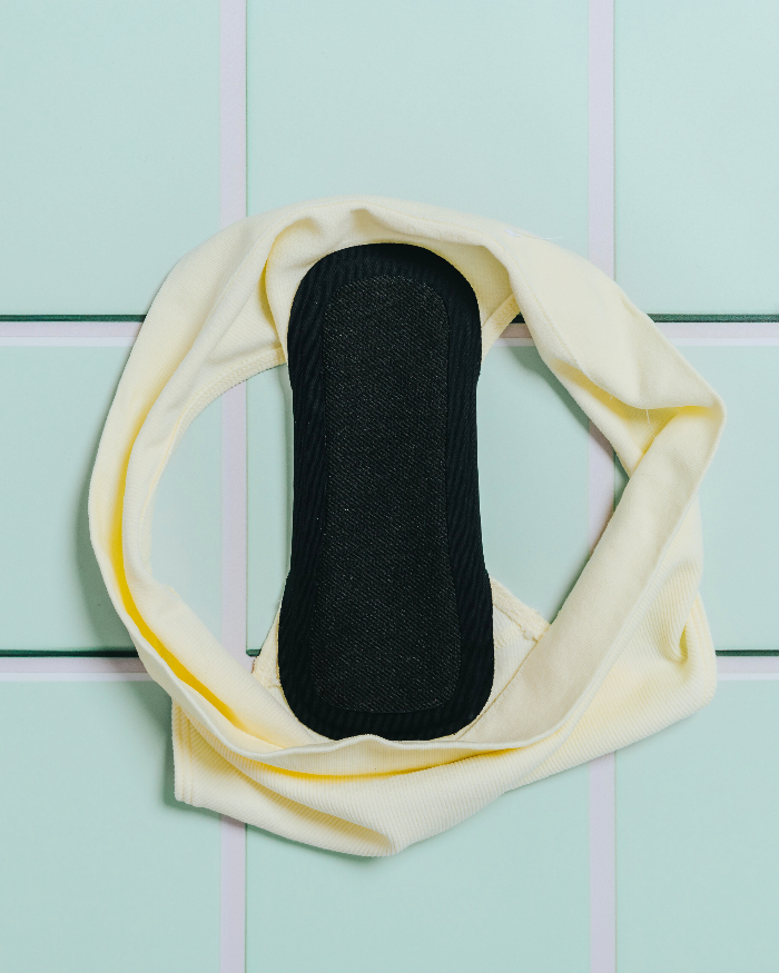 Black reusable panty liner attached to pair of pale yellow panties on a mint green tiled background  